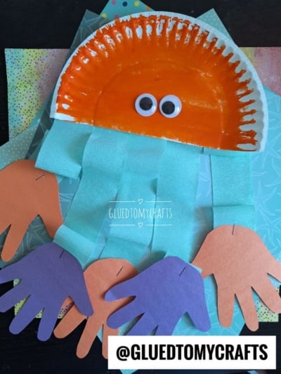 A octopus craft made with an orange-painted paper plate, googly eyes, blue crepe paper tentacles, and purple and orange handprint cutouts—fun inspiration for a paper plate octopus project. Background features layered patterned paper.