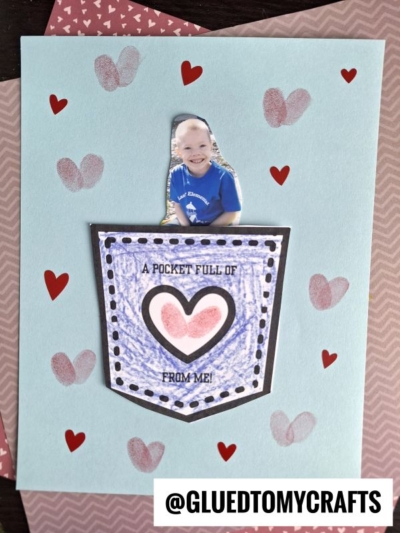 A handmade card featuring a paper pocket full of love, holding a cutout photo of a smiling child. The pocket says, “A pocket full of love from me!” The card is beautifully decorated with red and pink hearts. @GLUEDTOMYCRAFTS is at the bottom.