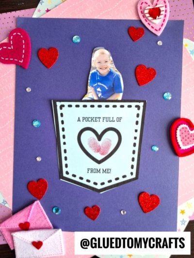 A handmade card featuring a paper pocket full of love, holding a cutout photo of a smiling child and surrounded by felt and sequin hearts. A label on the pocket reads, A pocket full of love from me! The username @GLUEDTOMYCRAFTS is at the bottom.