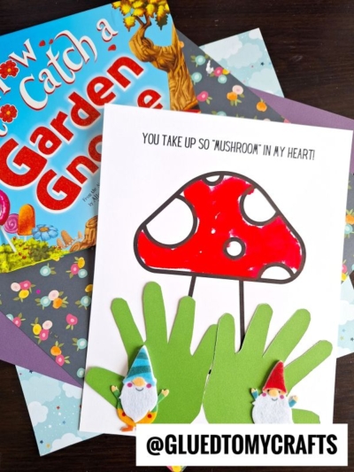 A handmade card with a red handprint mushroom, two gnome cutouts, and green handprint grass sits on floral paper. A book titled How to Catch a Garden Gnome is partially visible in the background.