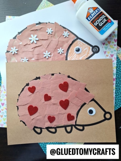 Two torn paper hedgehog crafts with googly eyes and glue. One features white flower shapes on white paper, while the other has red hearts on brown paper. A bottle of Elmer’s glue sits beside these adorable creations.