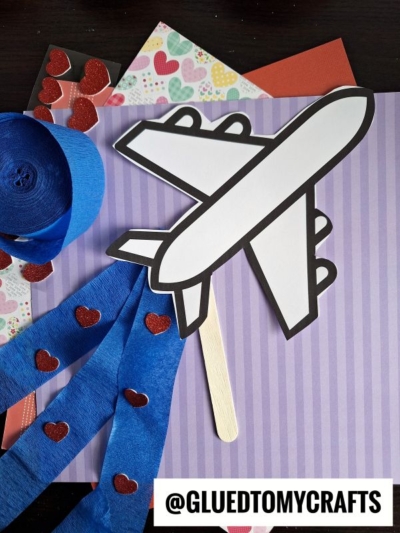 A crepe paper airplane craft with a popsicle stick handle and blue streamers adorned with red heart stickers, placed on striped and heart-patterned paper. The handle has the text @GLUETOMYCRAFTS printed next to it.