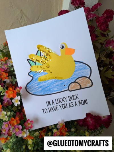 A handmade card featuring a handprint lucky duck with glitter on a pond, surrounded by flowers. The text reads: IM A LUCKY DUCK TO HAVE YOU AS A MOM! The @GLUEDTOMYCRAFTS handle is at the bottom.