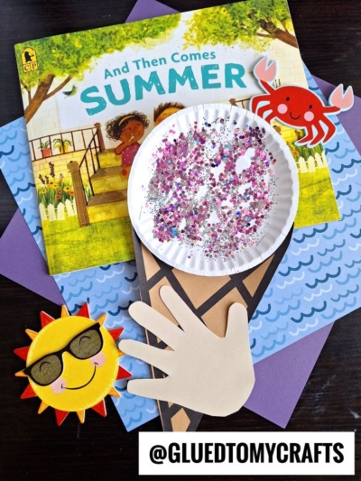 A children’s book titled “And Then Comes Summer” lies on a table, surrounded by colorful paper, a smiling sun, red crab, and a glittery paper plate snow cone craft with a handprint “cone.” @GLUEDTOMYCRAFTS.
