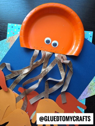 A paper plate octopus craft features an orange plate turned into a jellyfish, complete with googly eyes and shiny brown ribbon tentacles, displayed on colorful paper sheets. The Instagram handle @GLUEDTOMYCRAFTS appears in the corner.