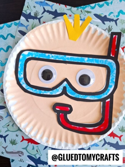 A paper plate snorkel craft featuring a snorkeler’s face, big googly eyes, a blue and red mask, and a yellow paper snorkel fin, all displayed on ocean-themed paper with sharks.