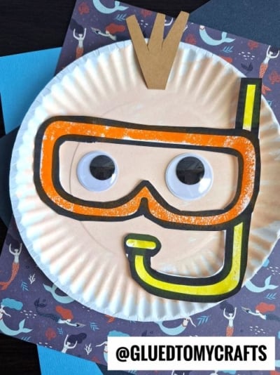 A paper plate snorkel craft features a snorkeler’s face with large googly eyes, a yellow and orange snorkel mask, and a brown paper hair piece, all set against a charming mermaid-themed background.