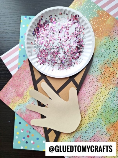 A paper plate snow cone with pink and silver glitter represents ice cream on a paper cone, held by a cut-out paper hand; colorful patterned papers create a playful background. @GLUEDTOMYCRAFTS is written at the bottom.