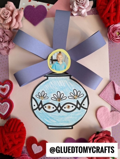 A handmade craft features a blue, vase-shaped drawing with large, 3-d paper flower vase petals and a photo of a child at the center, surrounded by decorative pink and red hearts and flowers on a polka dot background.