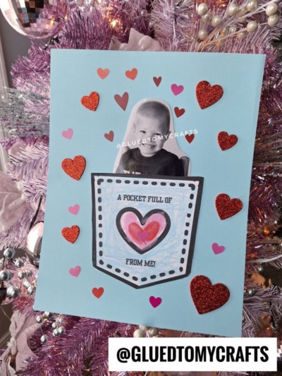 A handmade card with a photo of a smiling child above a paper pocket reading "A pocket full of love from me!" Red and pink hearts surround it, capturing the warmth of heartfelt affection. The background is a light blue card displayed on a pink Christmas tree, making this keepsake truly special.