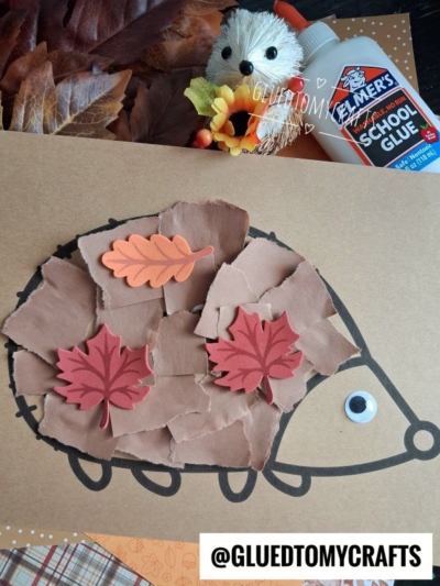 A torn paper hedgehog craft made of brown paper pieces and leaf cutouts on a drawn outline, with a googly eye. Glue, faux leaves, and a small hedgehog toy are nearby. @GLUEDTOMYCRAFTS is at the bottom.