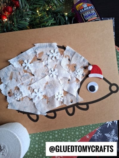 A torn paper hedgehog craft made from brown paper with white textured paper and snowflake cutouts, a googly eye, and a red Santa hat. Elmer’s glue, greenery, and assorted craft materials are visible nearby.