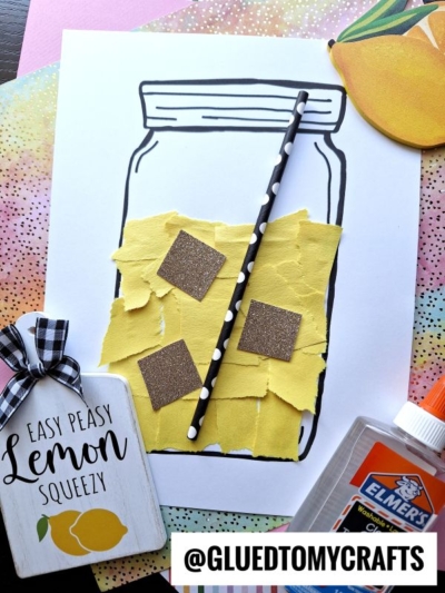 A craft project of a mason jar on paper, filled with yellow torn paper lemonade pieces, brown sparkly squares, and a black straw. Surrounded by craft supplies, a lemon tag, glue bottle, and colorful background paper.