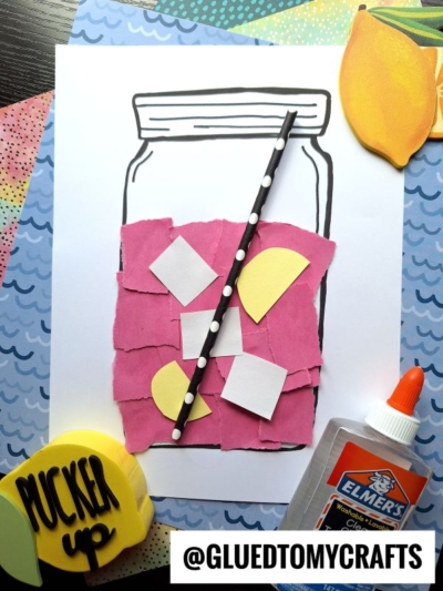 A paper craft of a mason jar filled with pink and white torn paper lemonade and yellow lemon slices, topped with a black polka-dot straw. Glue, a Pucker Up lemon decoration, and colorful patterned papers complete the scene.