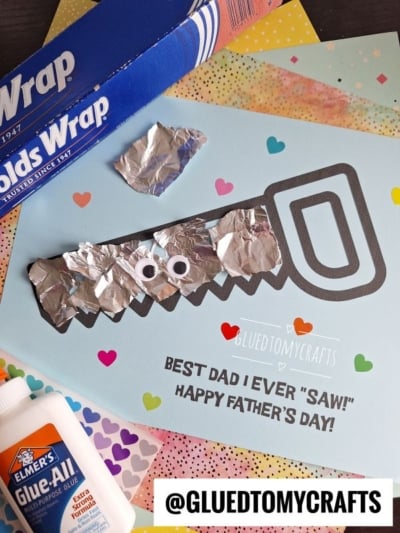 A homemade Father’s Day card shaped like a saw with googly eyes and aluminum foil, surrounded by colorful paper, glue, and a box of foil. The card reads, “Best dad I ever saw! Happy Father’s Day!”.