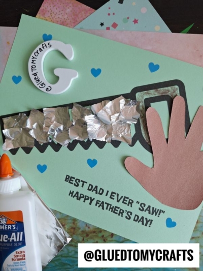 A handmade Father’s Day card features a unique saw shape and a charming handprint cutout, adorned with blue hearts and eye-catching foil accents. The heartfelt message reads: BEST DAD I EVER SAW! HAPPY FATHERS DAY! A glue bottle and the @GLUEDTOMYCRAFTS handle complete this creative tribute—perfect for celebrating the best dad you ever saw.
