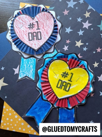 Two handmade #1 Dad Badge award ribbons with heart centers are displayed on star-patterned paper. One ribbon is red and blue, the other is yellow and red. The Instagram handle @GLUEDTOMYCRAFTS appears at the bottom.