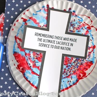 A paint splat Memorial Day craft featuring a white paper plate with red and blue paint, topped with a large cross that says, “Remembering those who made the ultimate sacrifice in service to our nation.”