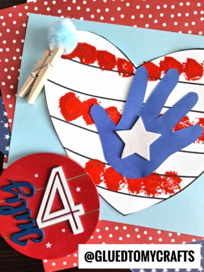 A patriotic July 4th craft featuring a heart with red paint stripes, a pom pom stamped flag handprint with a white star, a clothespin with blue pompom, and a red circle with July 4 on a polka-dot background.