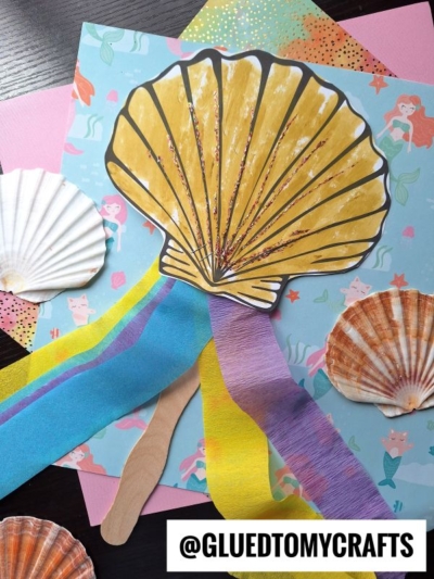A seashell wand craft made from yellow paper with glitter, colorful crepe paper streamers, and a popsicle stick, surrounded by real seashells on mermaid-themed paper. Text at the bottom reads @GLUEDTOMYCRAFTS.