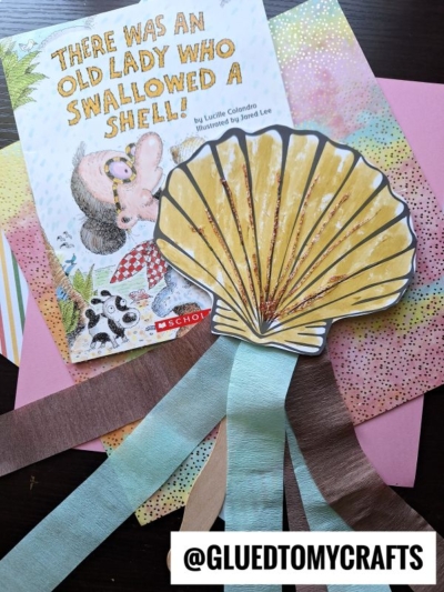A children’s book titled There Was an Old Lady Who Swallowed a Shell! is next to a colorful seashell wand craft with ribbon streamers and various vibrant sheets of paper in the background.