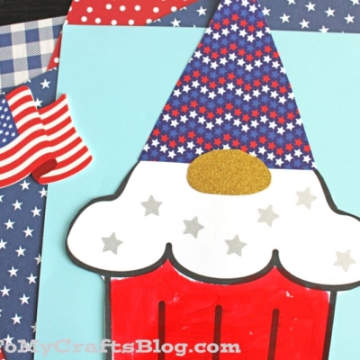 A patriotic cupcake gnome craft with a red base, white frosting with silver stars, and a blue hat covered in red, white, and blue stars, displayed on patterned paper with American flags.