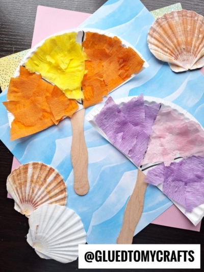 Colorful paper fans and a paper plate beach umbrella made from popsicle sticks and torn tissue paper in yellow, orange, and purple are displayed on blue paper with real seashells. A label at the bottom reads @GLUEDTOMYCRAFTS.