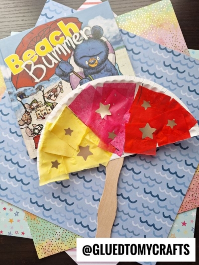 A colorful DIY paper plate beach umbrella craft sits on patterned paper next to a “Beach Bummer” children’s book. The umbrella is decorated with tissue paper and star cutouts, and @GLUEDTOMYCRAFTS is visible at the bottom.