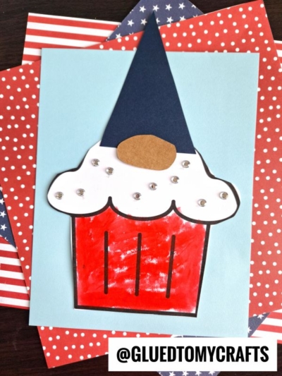 A handmade patriotic cupcake gnome craft features a red base, white icing with gem decorations, a blue pointed hat, and a brown oval nose. It sits on a blue sheet over red, white, and blue star-spangled and striped papers. @GLUEDTOMYCRAFTS.