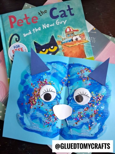 A children’s book titled Pete the Cat and the New Guy is shown with a blue paint splat cat face craft made from paint, paper, and colorful glitter, placed on top. The username @GLUEDTOMYCRAFTS appears at the bottom.