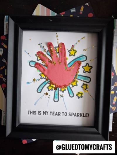 Framed artwork with a red handprint on white paper, decorated with glitter, yellow stars, and blue shapes. This year to sparkle keepsake features the text below that reads, THIS IS MY YEAR TO SPARKLE! Handle @GLUEDTOMYCRAFTS is at the bottom.