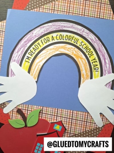 A handmade Back To School Rainbow craft with “I’m ready for a colorful school year!” written on it, featuring white hand cutouts on each side and a cheerful apple and plaid paper background. Instagram handle @GLUEDTOMYCRAFTS at the bottom.