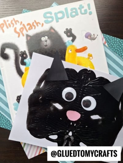 A children’s book with a black cat cover sits under a paint splat cat craft, made of black paint, paper ears, googly eyes, and a pink nose. The handle @GLUEDTOMYCRAFTS appears at the bottom.