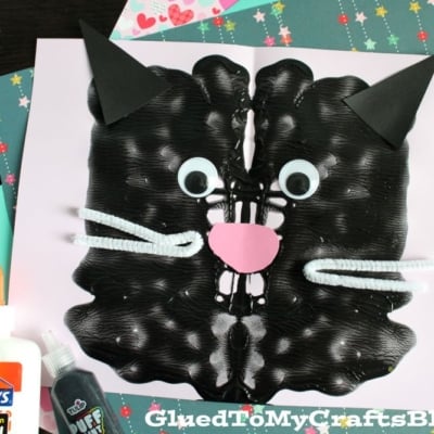A creative paint splat cat craft featuring a black paint splat shaped like a cat, with googly eyes, pipe cleaner whiskers, a pink paper nose, and black paper ear, surrounded by glue, glitter, and star-patterned paper.