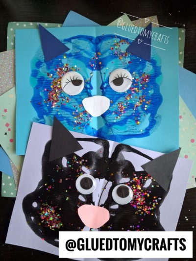 Two colorful paint splat cat art pieces made from paint, googly eyes, paper ears, and confetti are displayed on pastel paper. Each cat has a heart-shaped nose, and the username @gluedtomycrafts is visible on the image.