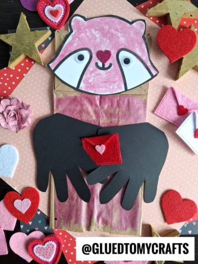 A paper bag raccoon craft made with colored paper and felt hearts. The raccoon holds a red felt envelope with black paper arms, surrounded by Valentine-themed stars and hearts. @GLUEDTOMYCRAFTS is at the bottom.