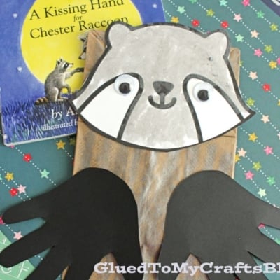 A paper bag raccoon craft with googly eyes and black paper hands is displayed on patterned paper beside the book A Kissing Hand for Chester Raccoon.