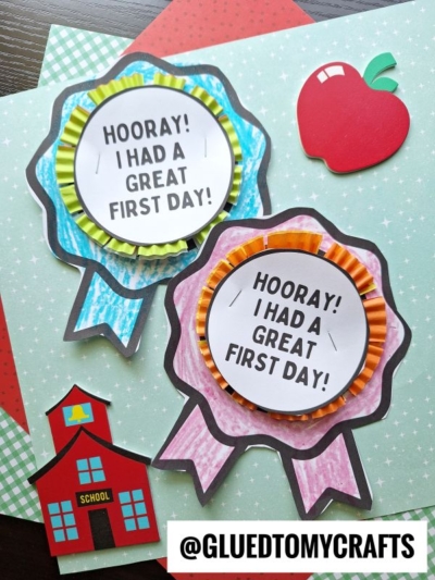 Two colorful paper ribbons with HOORAY! I HAD A GREAT FIRST DAY! text create a cheerful first day of school badge, set on a green star-patterned background with a red apple, small red schoolhouse, and @GLUEDTOMYCRAFTS at the bottom.