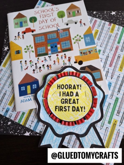 A children’s book titled “School’s First Day of School” sits on patterned paper with a colorful handmade first day of school badge that reads “Hooray! I had a great first day!” @GluedToMyCrafts is labeled at the bottom.