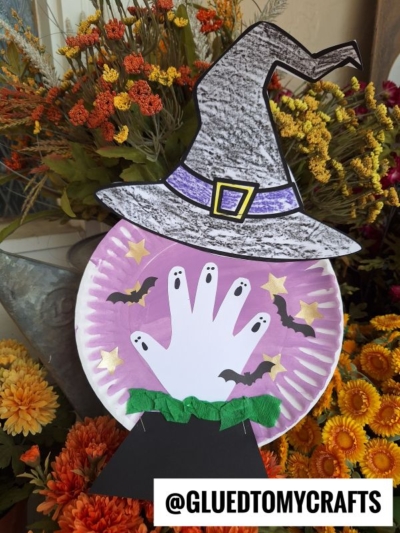 A Paper Plate Halloween Snow Globe craft featuring a purple background, a white handprint ghost, black bats, yellow stars, and a witch hat, all surrounded by colorful flowers. @GLUEDTOMYCRAFTS is written at the bottom.