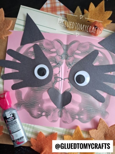 A pink paper with a butterfly inkblot, black paper shapes, and googly eyes forms a cat face inspired by the playful look of a paint splat raccoon. Fall leaves, plaid paper, and silver paint are nearby. @GLUEDTOMYCRAFTS is written on the image.