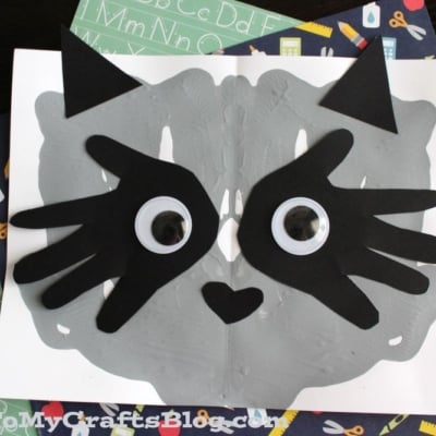 A paint splat raccoon art project features a gray splattered face, black paper ears and eye patches, big googly eyes, and a heart-shaped nose.