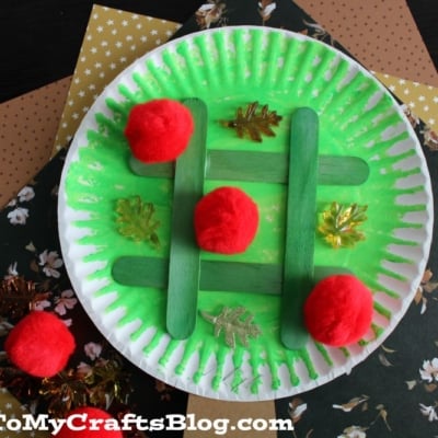 Paper Plate Apple Tree Tic Tac Toe Game Craft Idea For Kids To Make - A colorful paper plate craft designed as a tree with red pom-poms and green decorations.