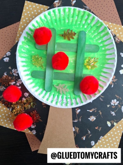 A paper plate painted green with green popsicle sticks, red pom-poms, and leaf-shaped decorations on it, resembling an apple tree tic tac toe craft, sits on patterned paper backgrounds.