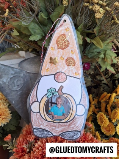 A charming fall gnome photo ornament paper craft featuring a child’s photo in the center, autumn leaves, and pumpkin decorations, all surrounded by vibrant fall flowers. The text @GLUEDTOMYCRAFTS is displayed at the bottom.