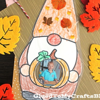 A fall-themed gnome craft made with paper, featuring a child's photo in a pumpkin, autumn leaves, and colored decorations.