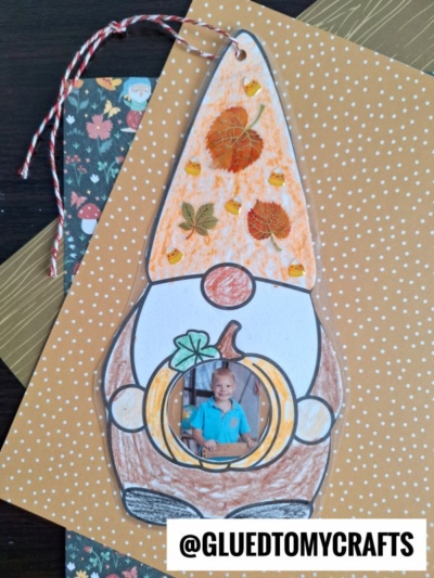 A charming fall gnome photo ornament craft featuring a paper gnome decorated with leaves, gems, and a child’s photo nestled inside a pumpkin shape, all displayed on layered patterned papers. @GLUEDTOMYCRAFTS is written at the bottom.