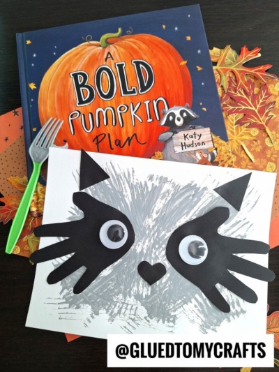 Childrens book A Bold Pumpkin Plan with a delightful fork painted raccoon art project, featuring a raccoon face created with fork painting, paper cut-out ears, and large googly eyes, displayed on a dark surface with autumn leaves.
