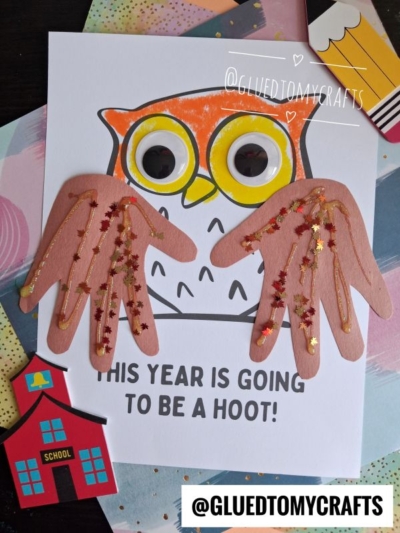 A vibrant handprint owl craft with googly eyes and wings made from brown paper sits atop a card that reads, This year is going to be a hoot! Surrounding the handprint owl are school-themed paper cutouts and sparkling glitter accents.