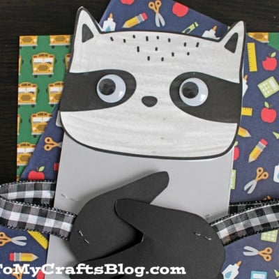A handprint raccoon craft with googly eyes and a plaid ribbon sits on colorful, school-themed papers featuring icons like school buses, apples, and scissors. The image is watermarked with GluedToMyCraftsBlog.com.